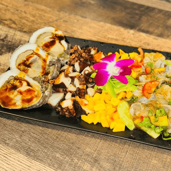 A black platter of oysters, octopus, mango, and shrimp ceviche, garnished with a purple orchid.