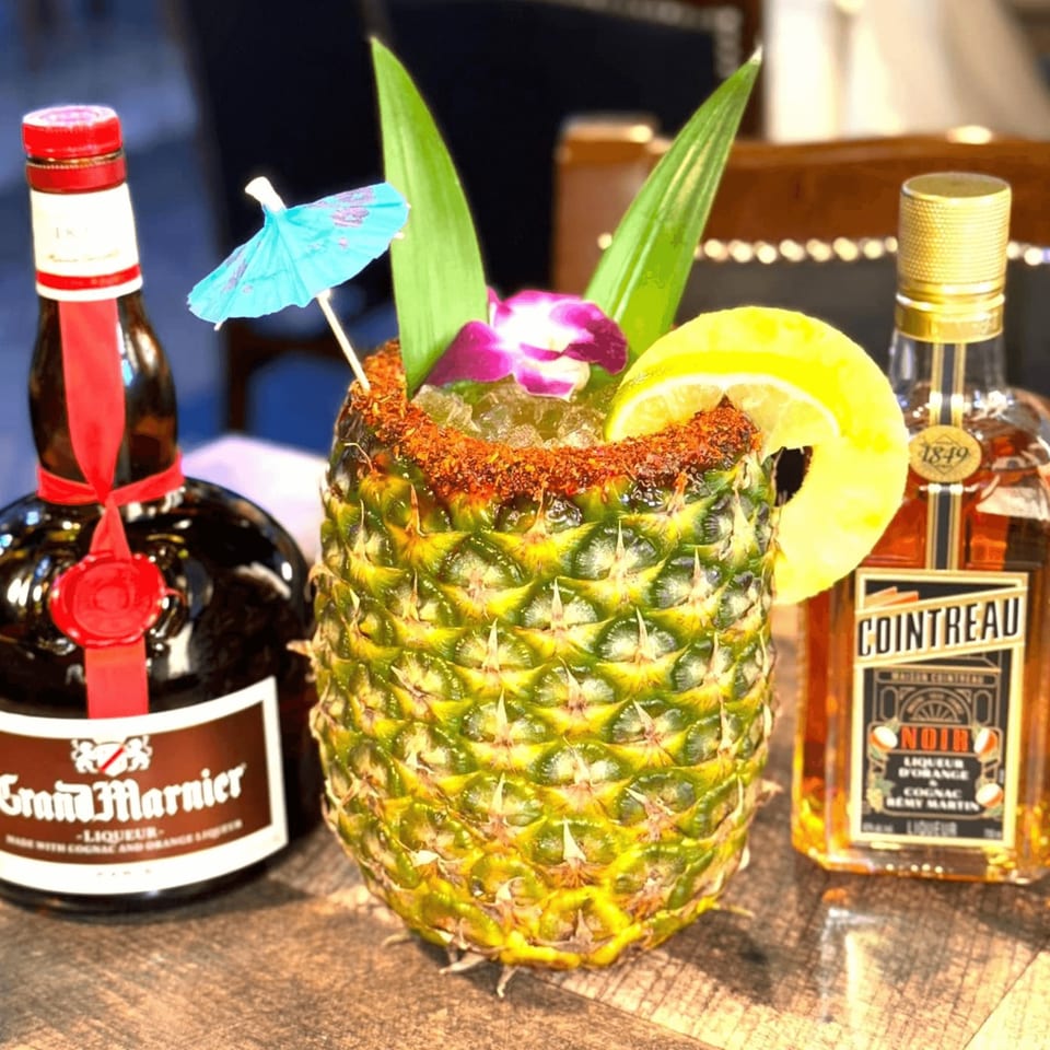 A pineapple cocktail with a lime slice, purple orchid, and umbrella, flanked by Grand Marnier and Cointreau Noir bottles.