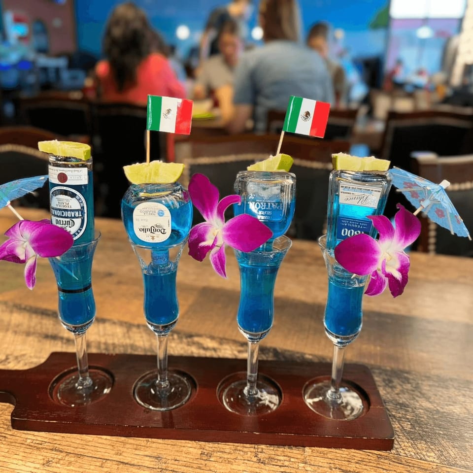 Four blue tequila shots with miniature bottles, lime wedges, orchids, and Mexican flags on a wooden tray.