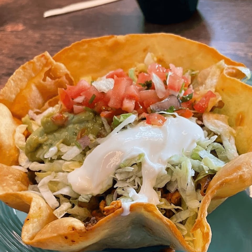A taco salad in a crispy tortilla bowl, topped with guacamole, pico de gallo, and sour cream.