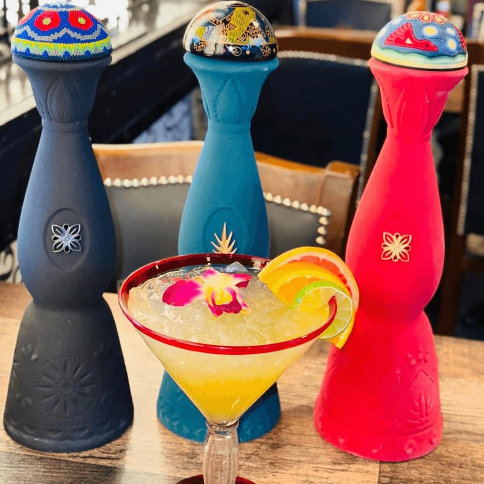 A vibrant margarita with a flower and citrus garnish, surrounded by three colorful, decorative tequila bottles.