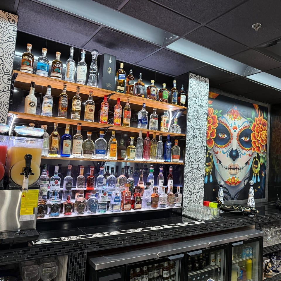 A well-stocked bar at La Plaza Fiesta with numerous liquor bottles and a vibrant Day of the Dead mural.
