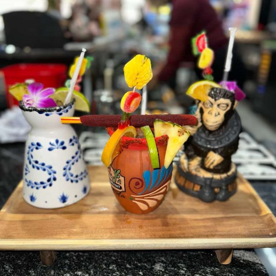 Three colorful, garnished Mexican drinks in unique ceramic mugs on a wooden tray.