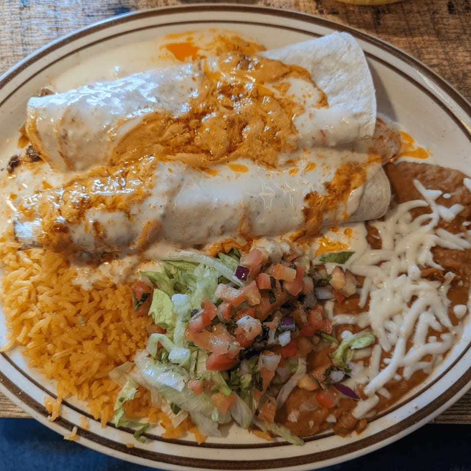Two burritos covered in white and red sauce, with rice, refried beans, and pico de gallo.