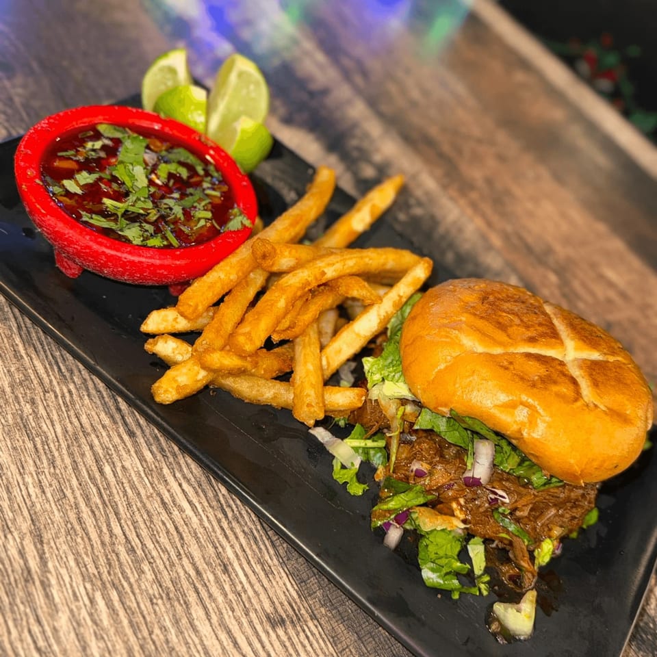 Birria burger with fries and a side of consommé with lime wedges on a black plate.